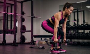 A middle-aged woman in black and pink athletic wear performs a Romanian Deadlift in a commercial gym.